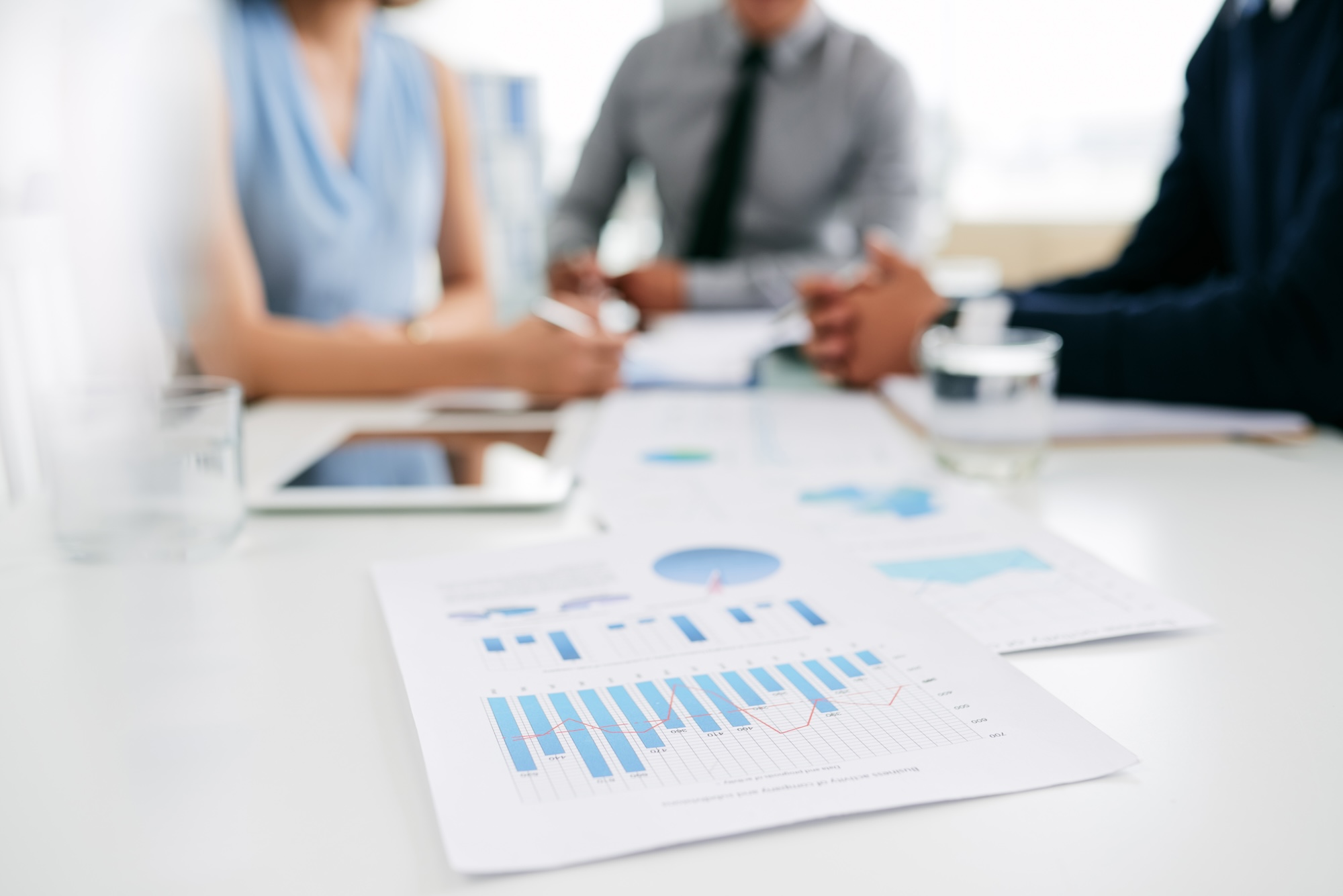 Charts and graphs on paper in the foreground and three individuals discussing costs in the background